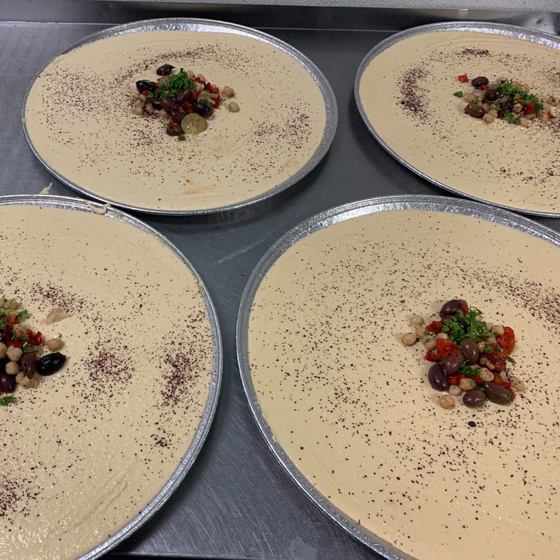 Four large round trays of hummus, garnished with olives, chickpeas, red peppers, and parsley.